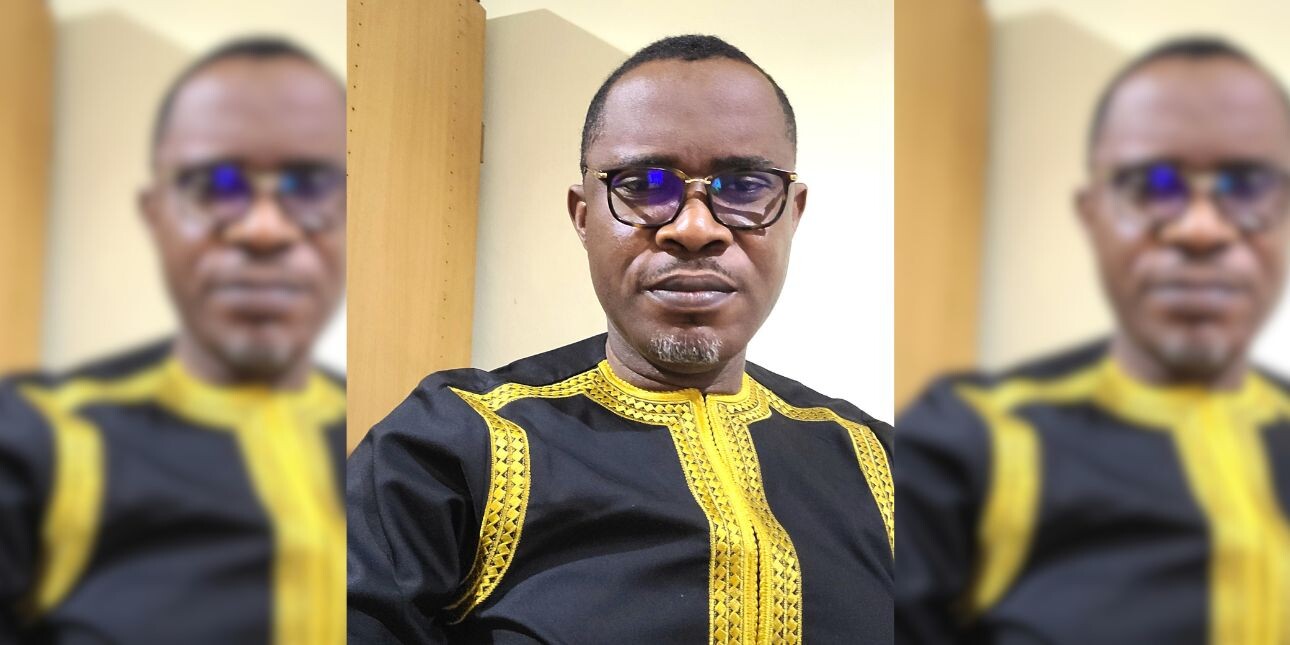 Portrait of Ezenwa Opara, a shaven haired black man with glasses and a goatee beard wearing a navy top with gold embroidery